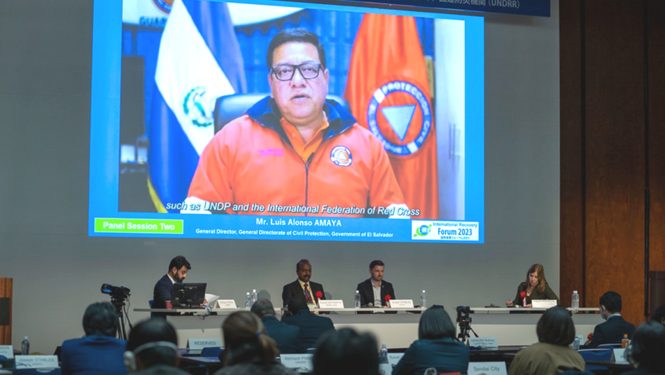 A virtual speaker speaks to a conference room full of guests on a large screen, dressed in emergency responder wear, at the International Recovery Forum 2019, run by UNDRR's International Recovery Platform.