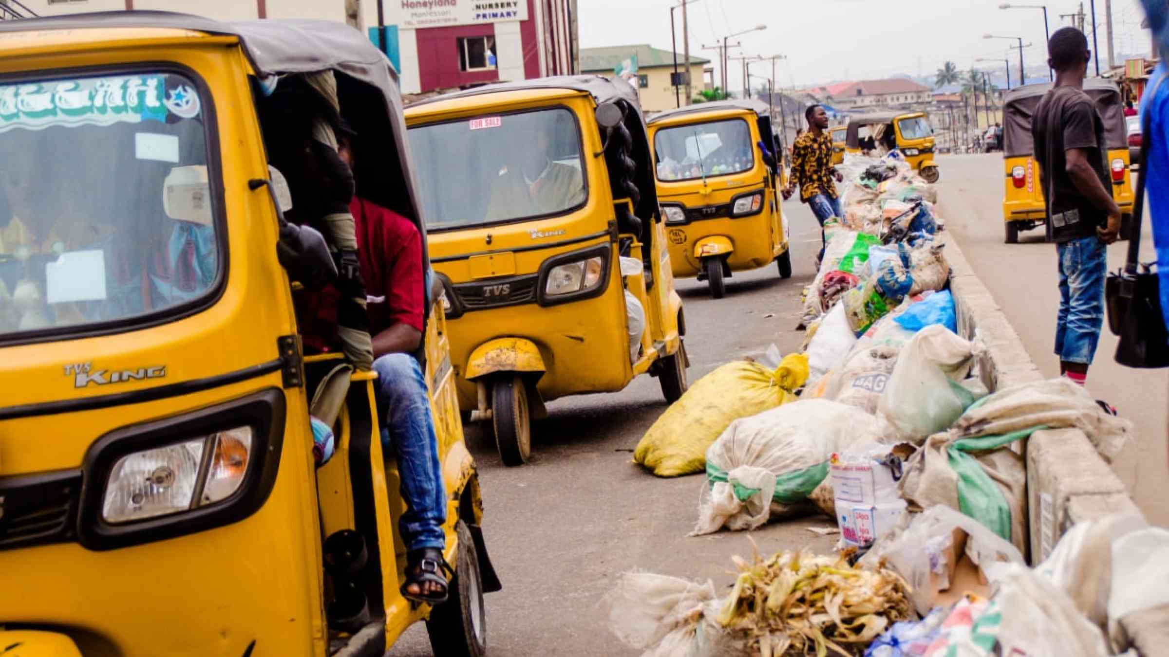 Waste laying on the streets in Nigeria. Yellow tricycles pass the waste along the road.