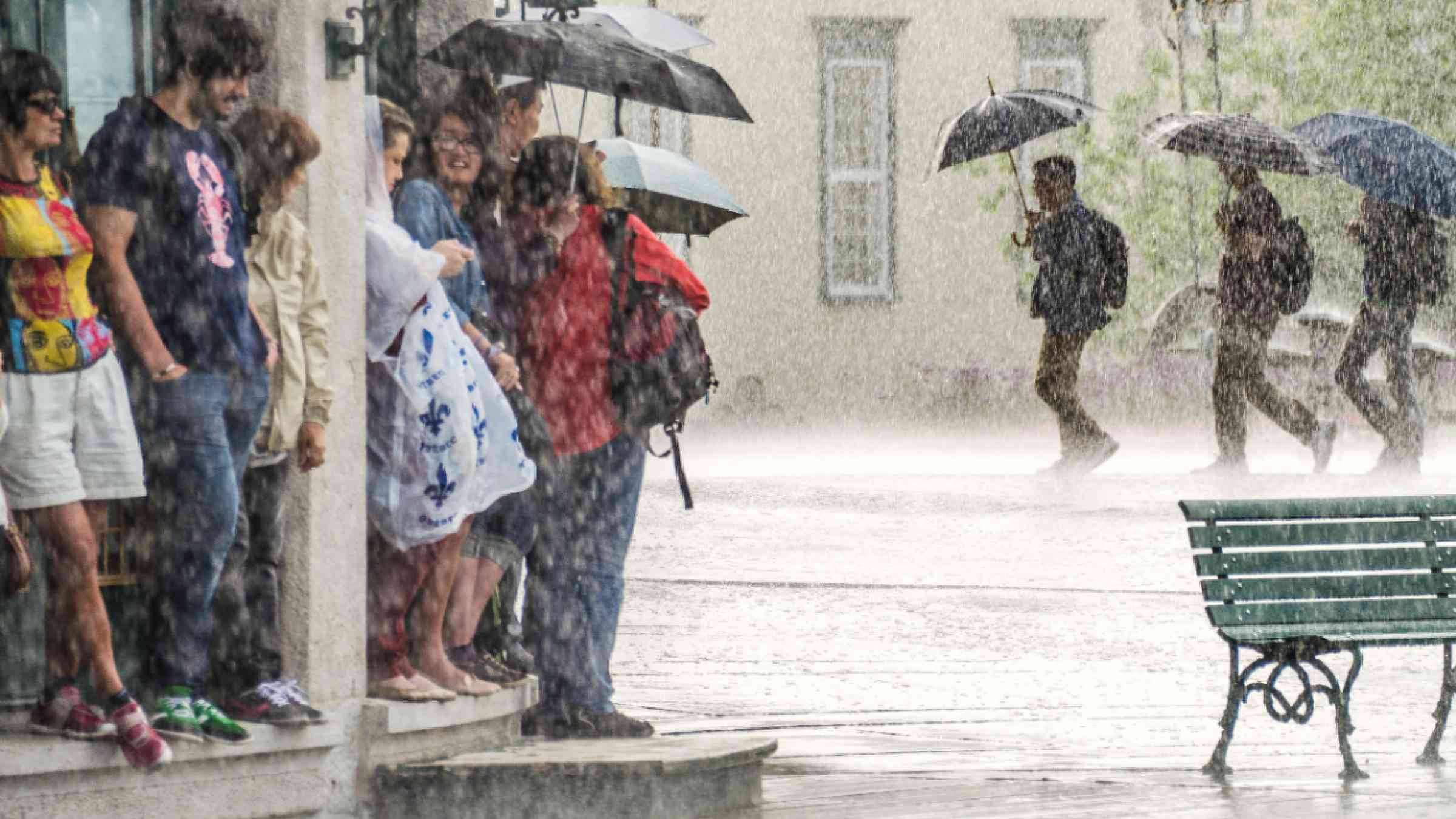A group of people hide from heavy rain under a building while three persons in the background walk with umbrellas