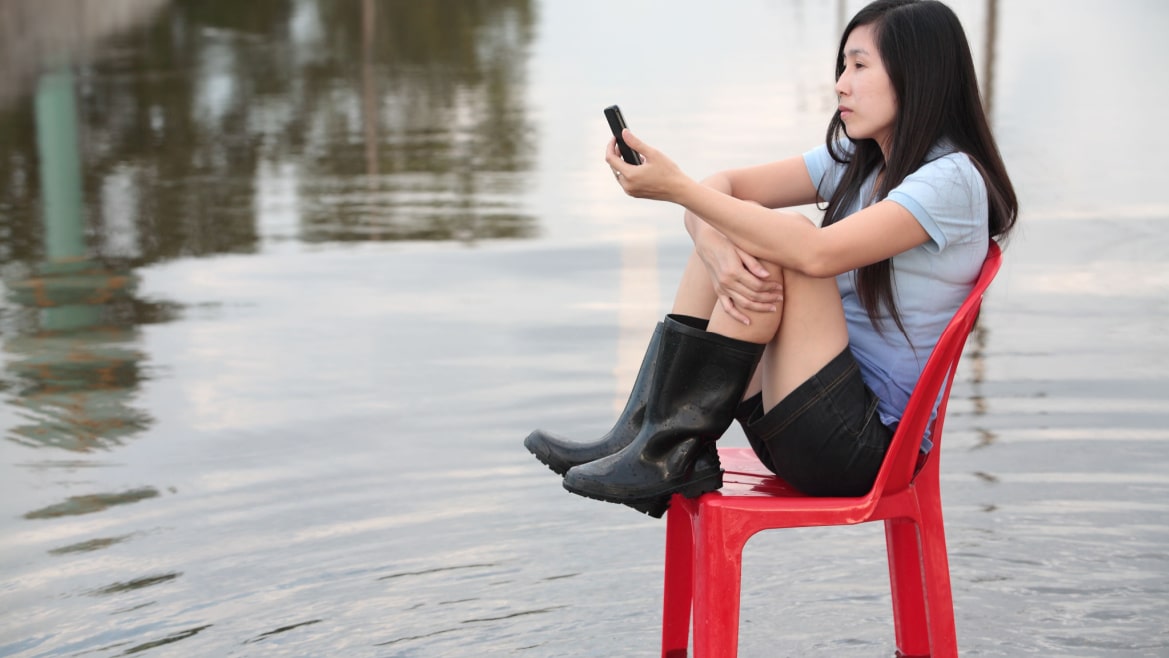 Asian woman sitting on a red chair looking at her phone in flooded street.