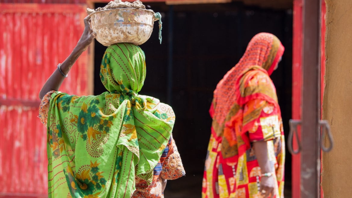 Women dressed in traditional closes entering their house in the Chad. One is carrying a basket on her head.