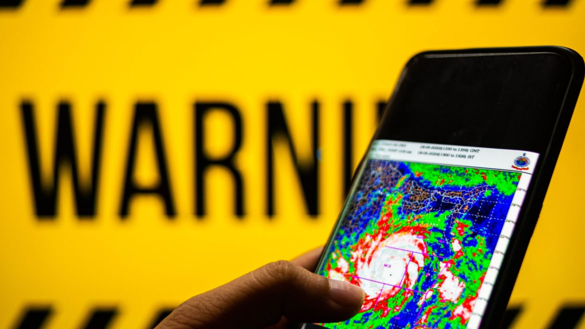Photograph of a mobile phone with cyclone moving towards the coast shown. The background is yellow with warning sign.