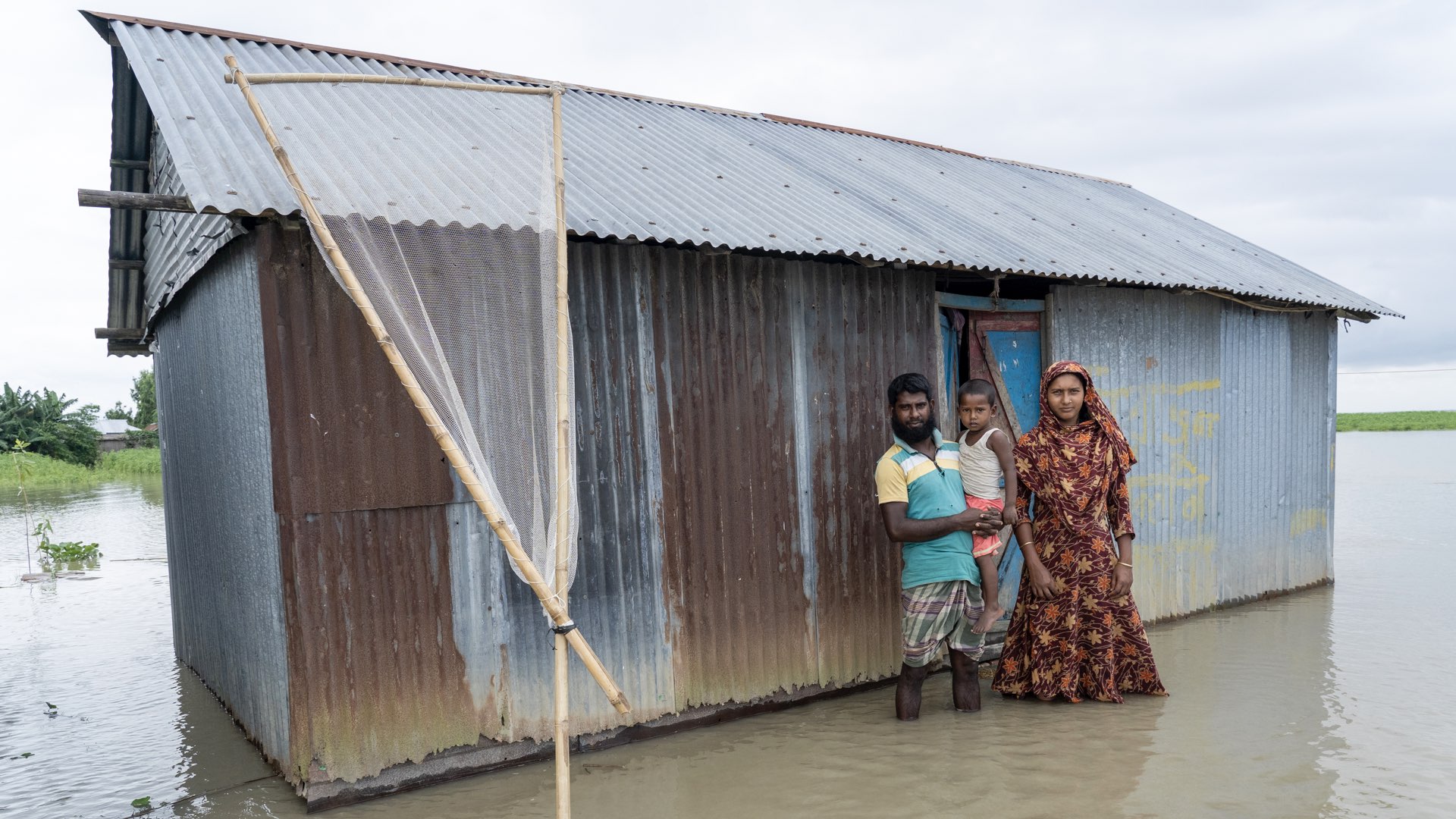 Family of three in Bangladesh posing in front of their house amidst floods
