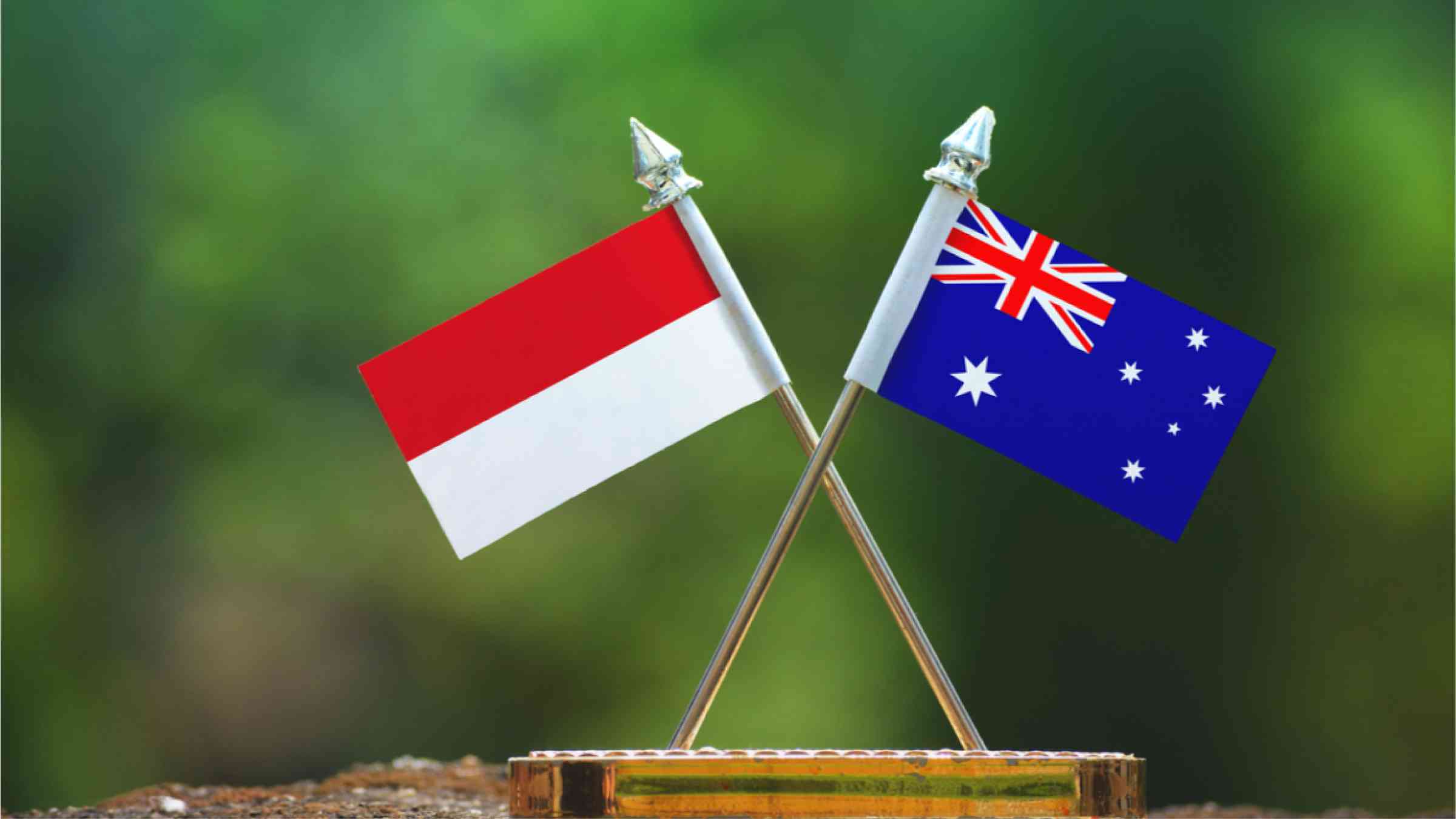 This image shows the two flags of Australia and Indonesia next to each other in front of blurred green nature.