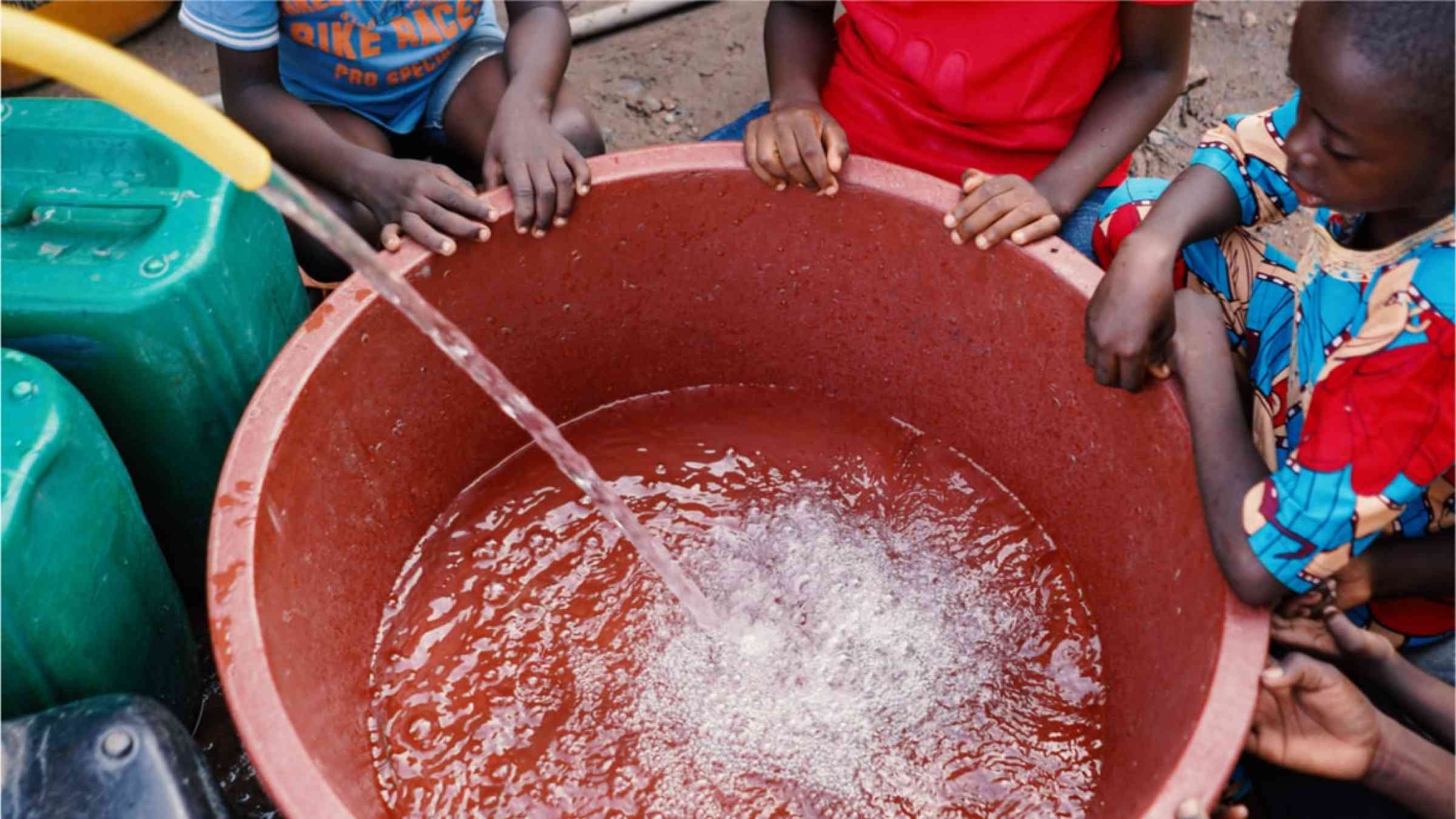 Three children gathering around a large bucket that is being filled with fresh drinking water.