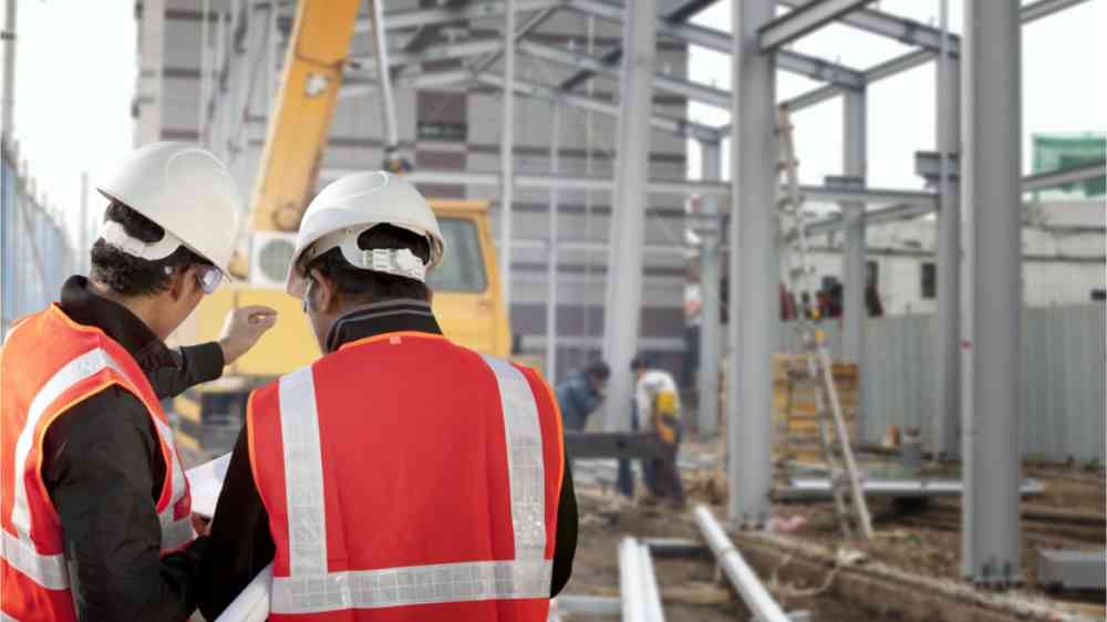 Two construction workers looking at a printed plan and building that is currently being built.