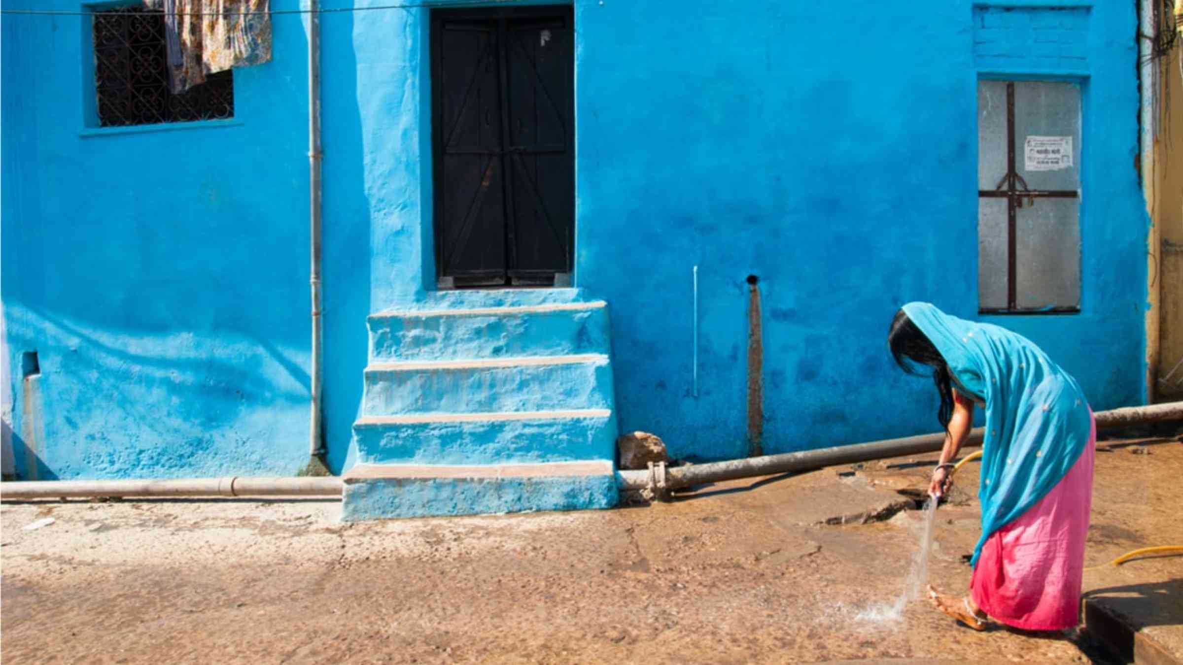 An Indian woman sparkling water on to the street in front of her light blue house.