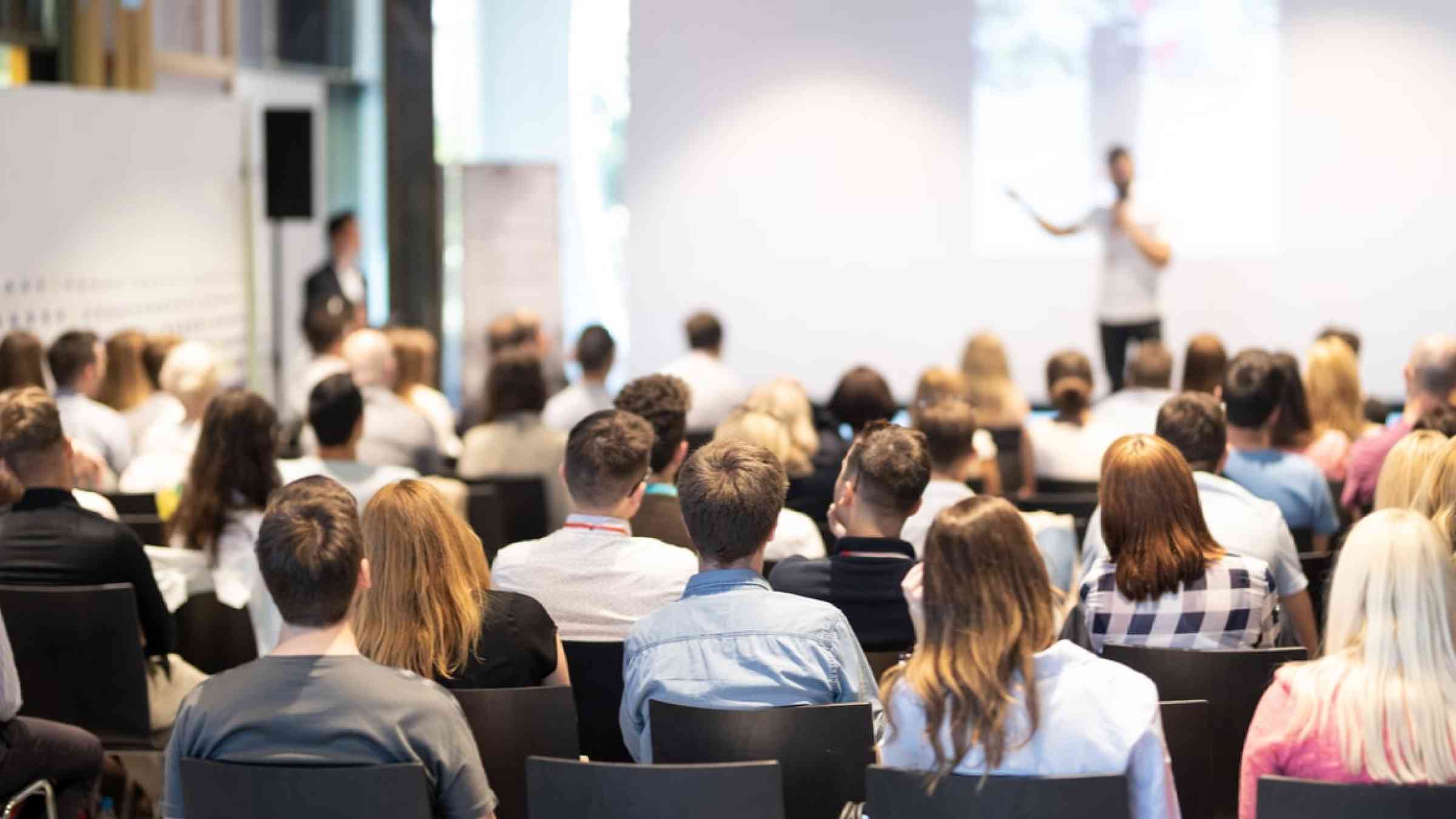 A conference room full of people with a person in the front holding a presentation.