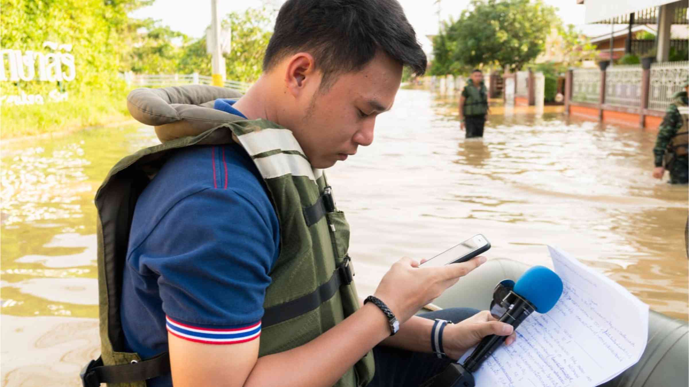 Journalist writing notes in his phone and on paper while holding his microphone in a heavy flooded area in Thailand.