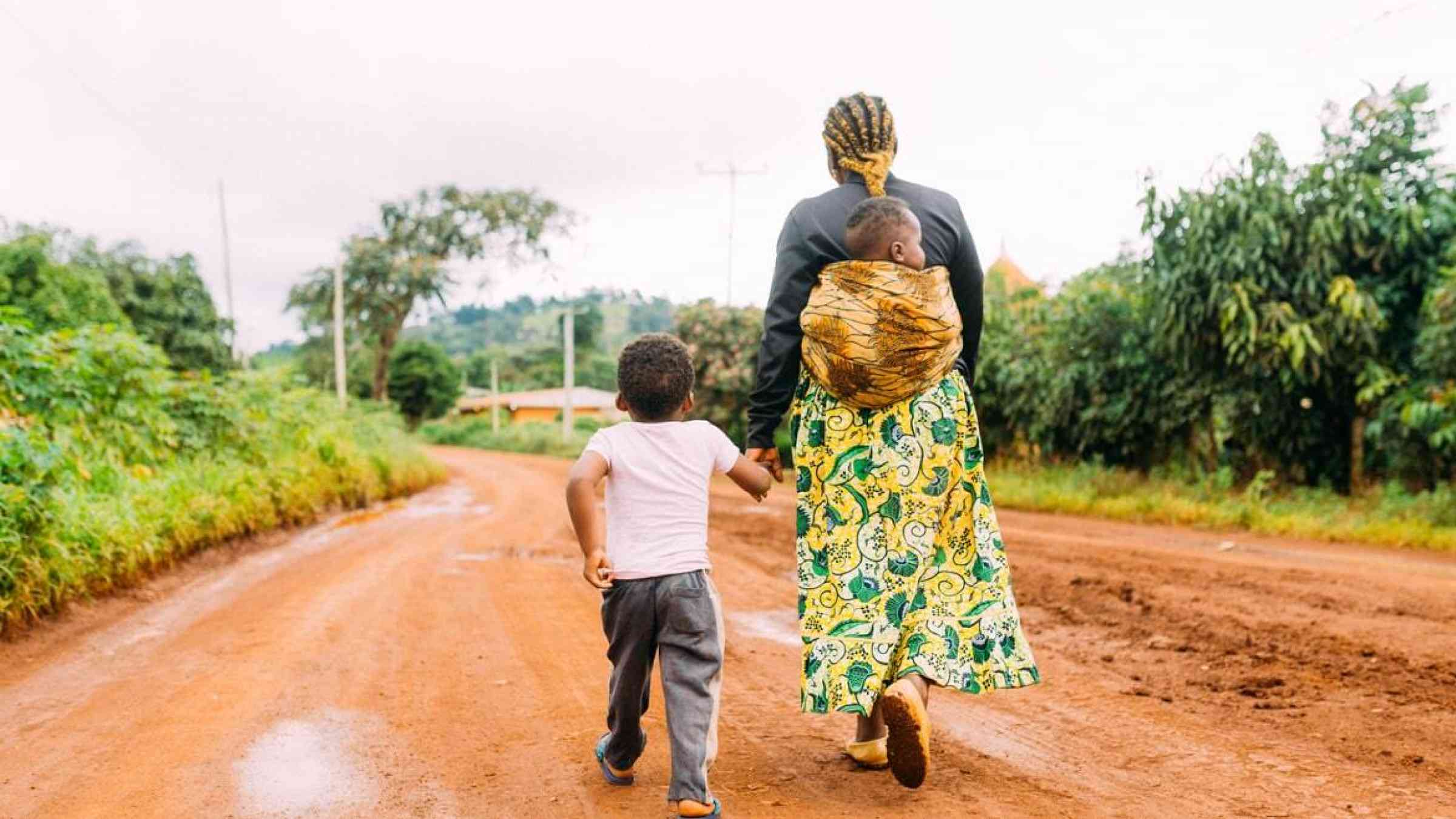 A woman carrying a baby and holding the hand of a boy, walking in a muddy street