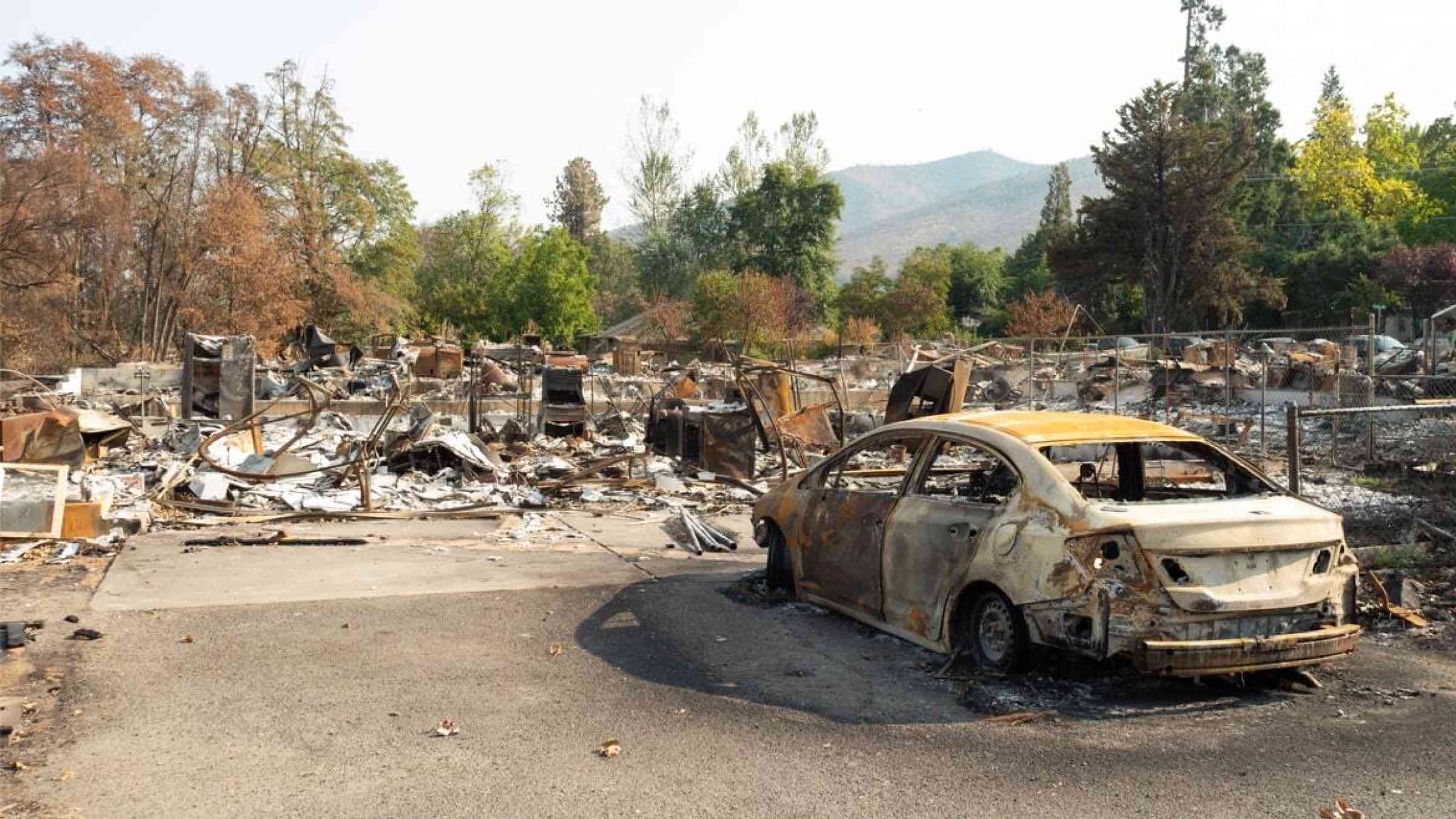 A burned out car sits among the remains of a burned neighborhood from wildfires, healthy vegetation in the background