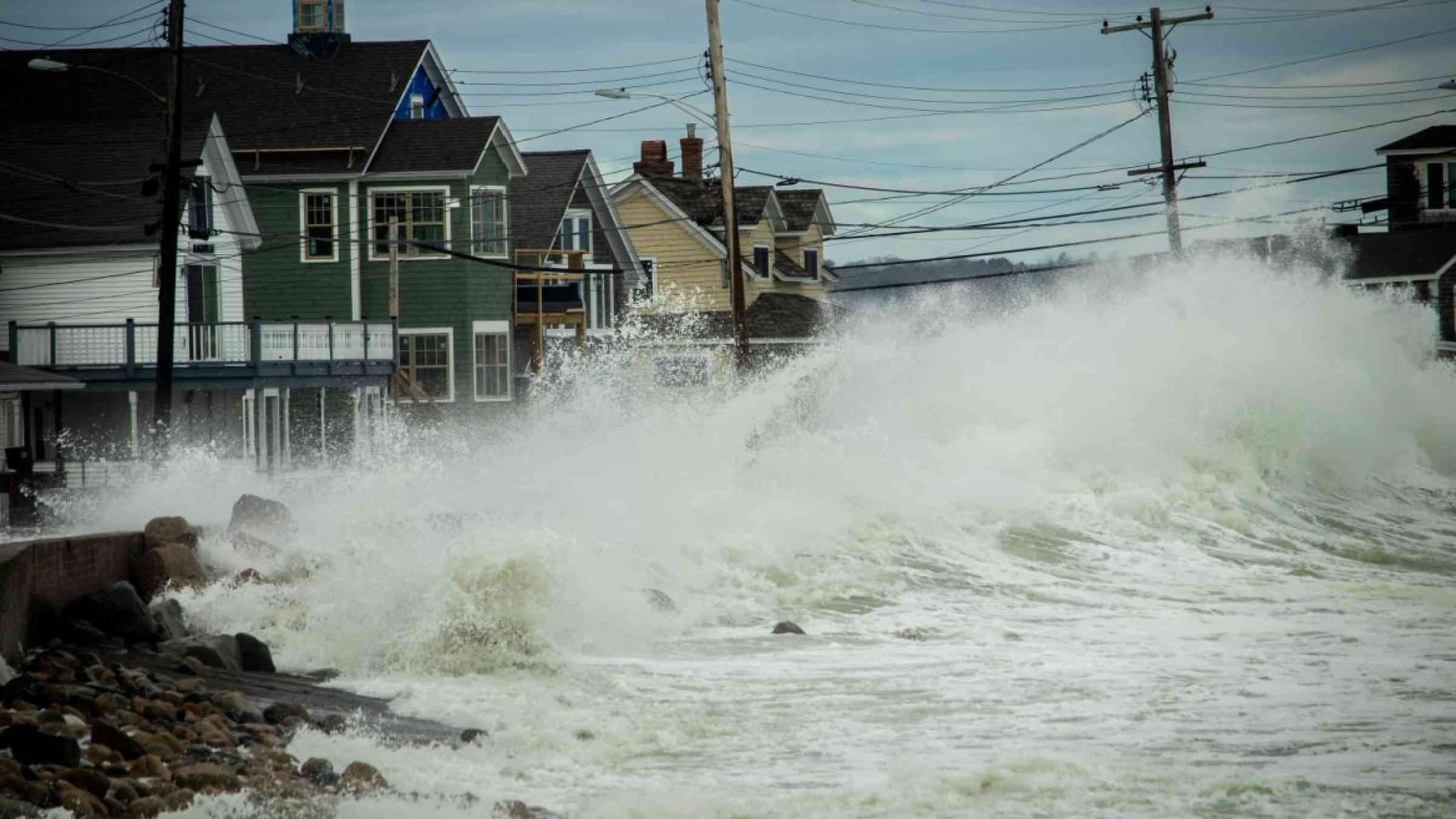 A storm ocean wave crashes over the road and floods coastal houses.