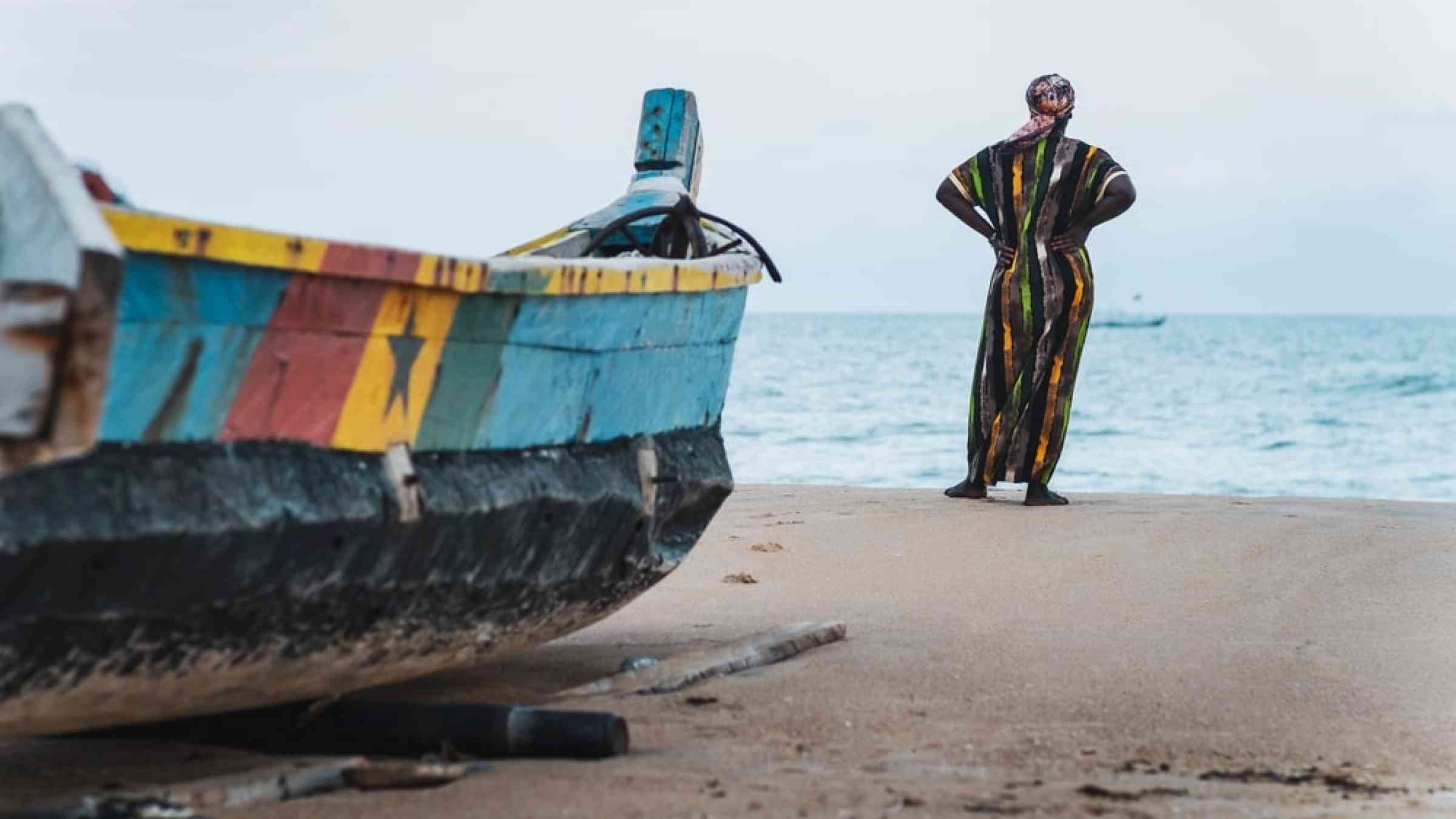 African woman in long suit standing by the shore and looking out over the sea in Keta Ghana, West Africa.