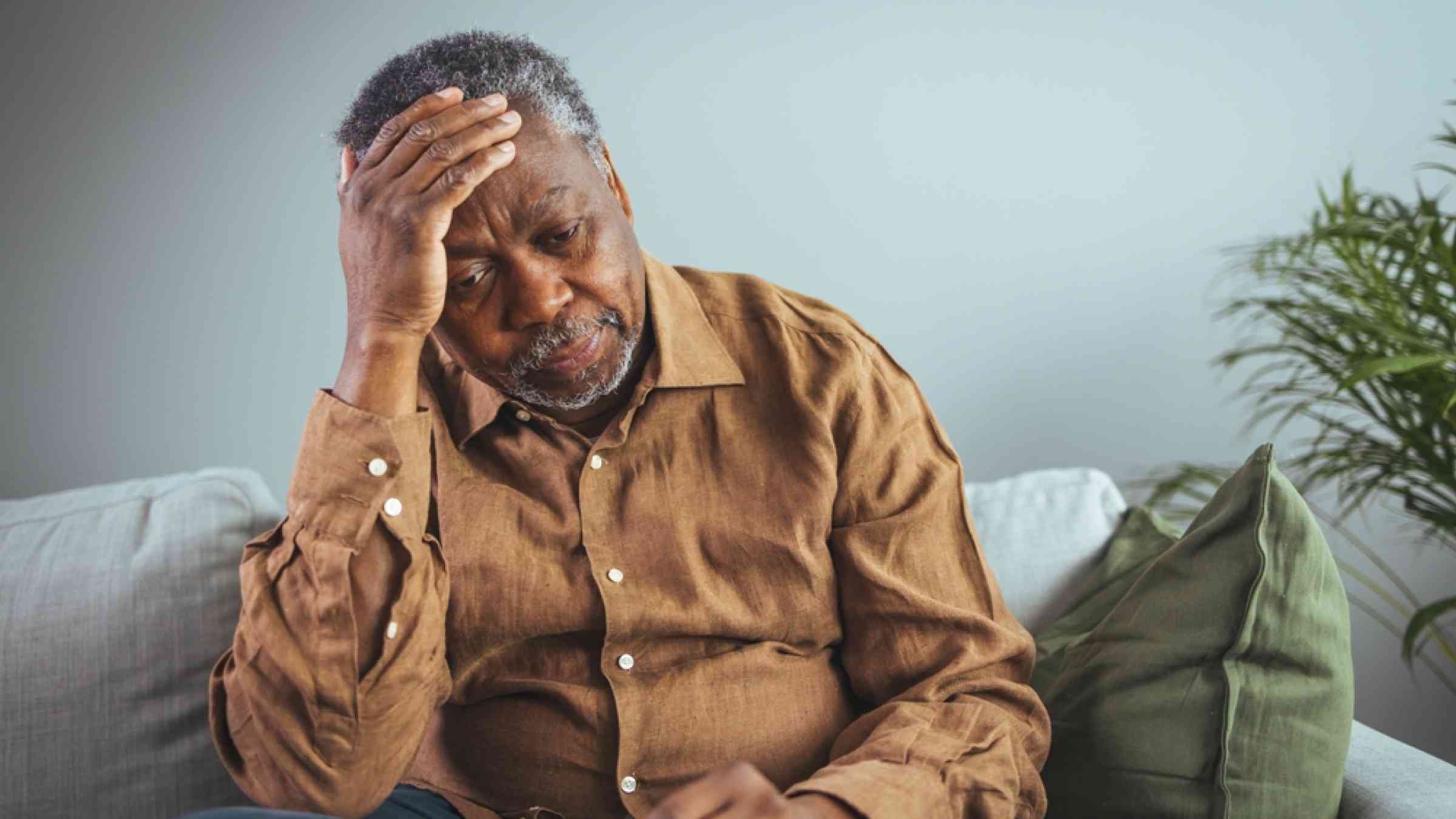 A Black adult man has a headache. He sits and holds his hands on his head in a dark room.