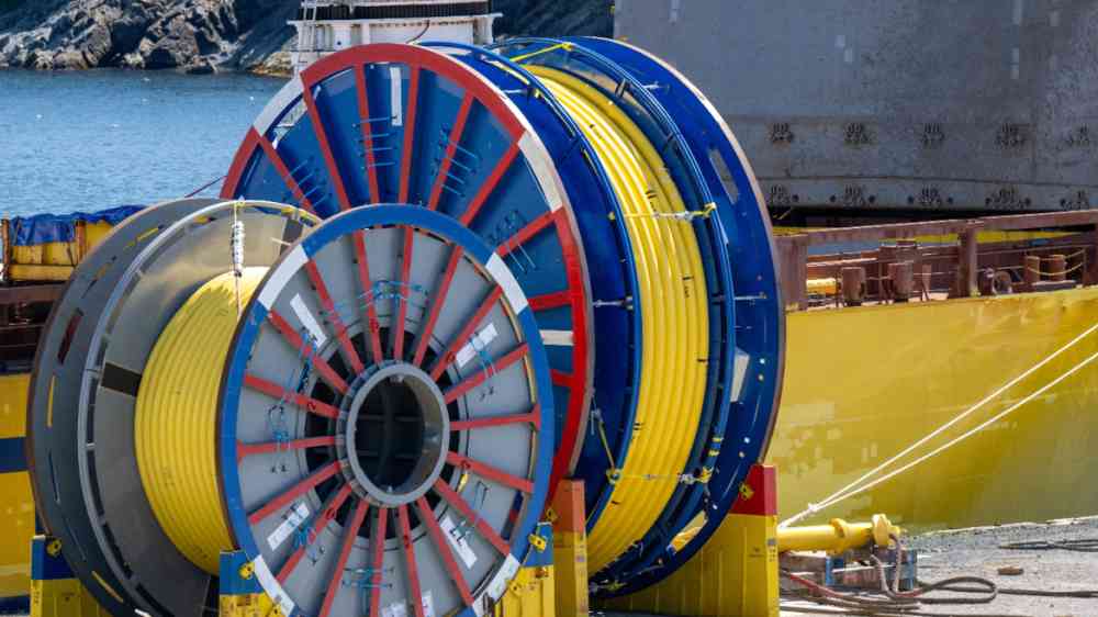 Bulk sub-sea industrial glass fiber optic cable on a metal spool on a ship's stand. The yellow data line is coiled around a black reel in a storage yard. Internet communications spool storage yard.