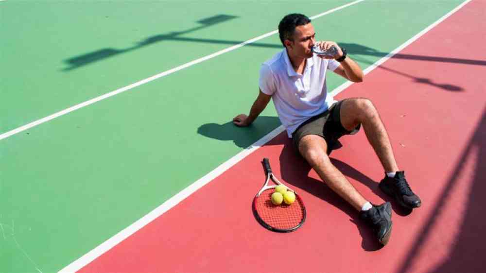  Young Latin male tennis player resting and drinking water on tennis court in sunny hot day at summer