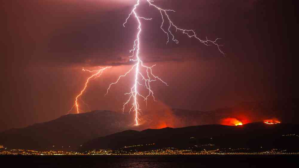 Lightning illuminates the night sky over a mountainous landscape with distant wildfires and city lights.