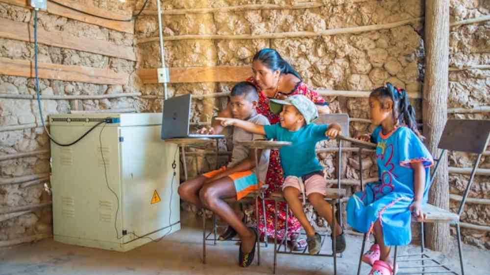 Teacher helps children learn on laptop in rustic room.