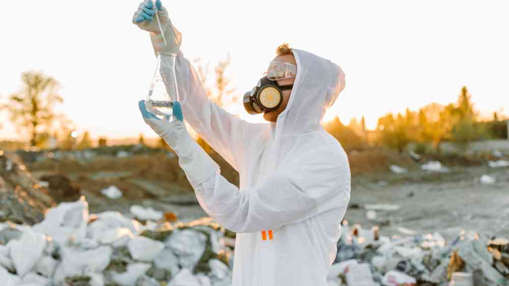 Scientist in protective gear collecting samples at a waste site during sunset.