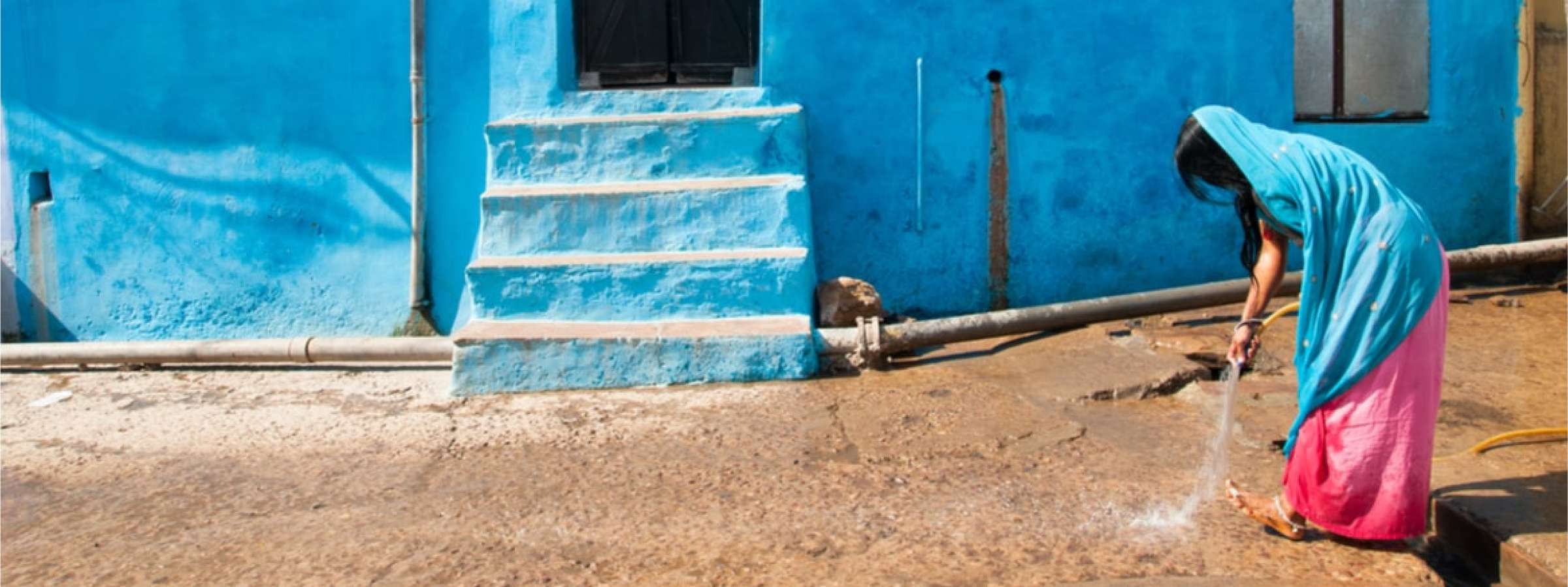 An Indian woman sparkling water on to the street in front of her light blue house.